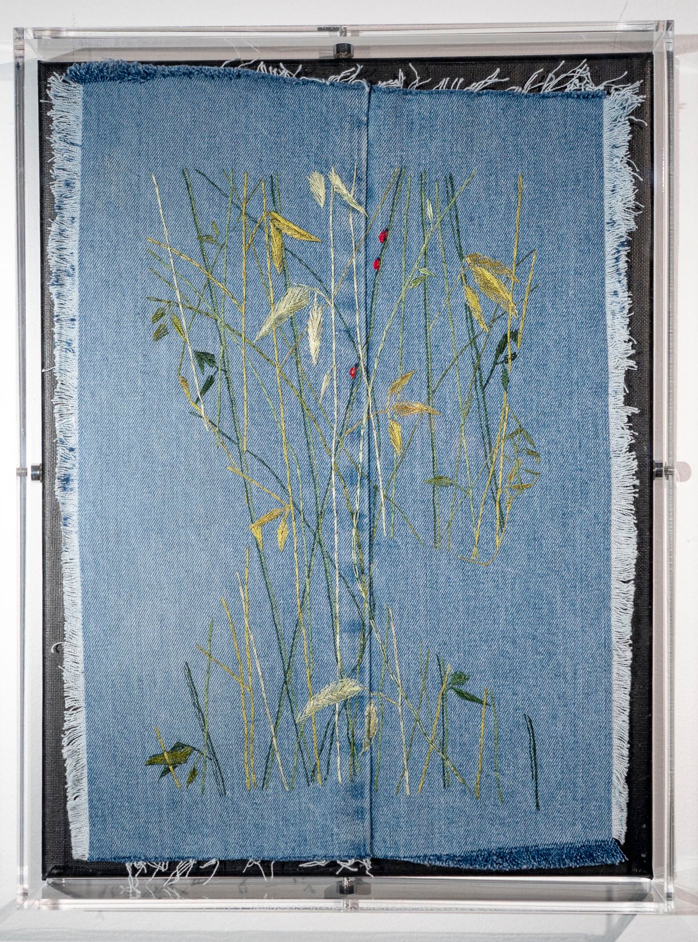 On frayed blue denim, a face is revealed in the negative by seeing the outline made up of a growth of bamboo shoots dotted with three ladybugs, done in glossy threads.