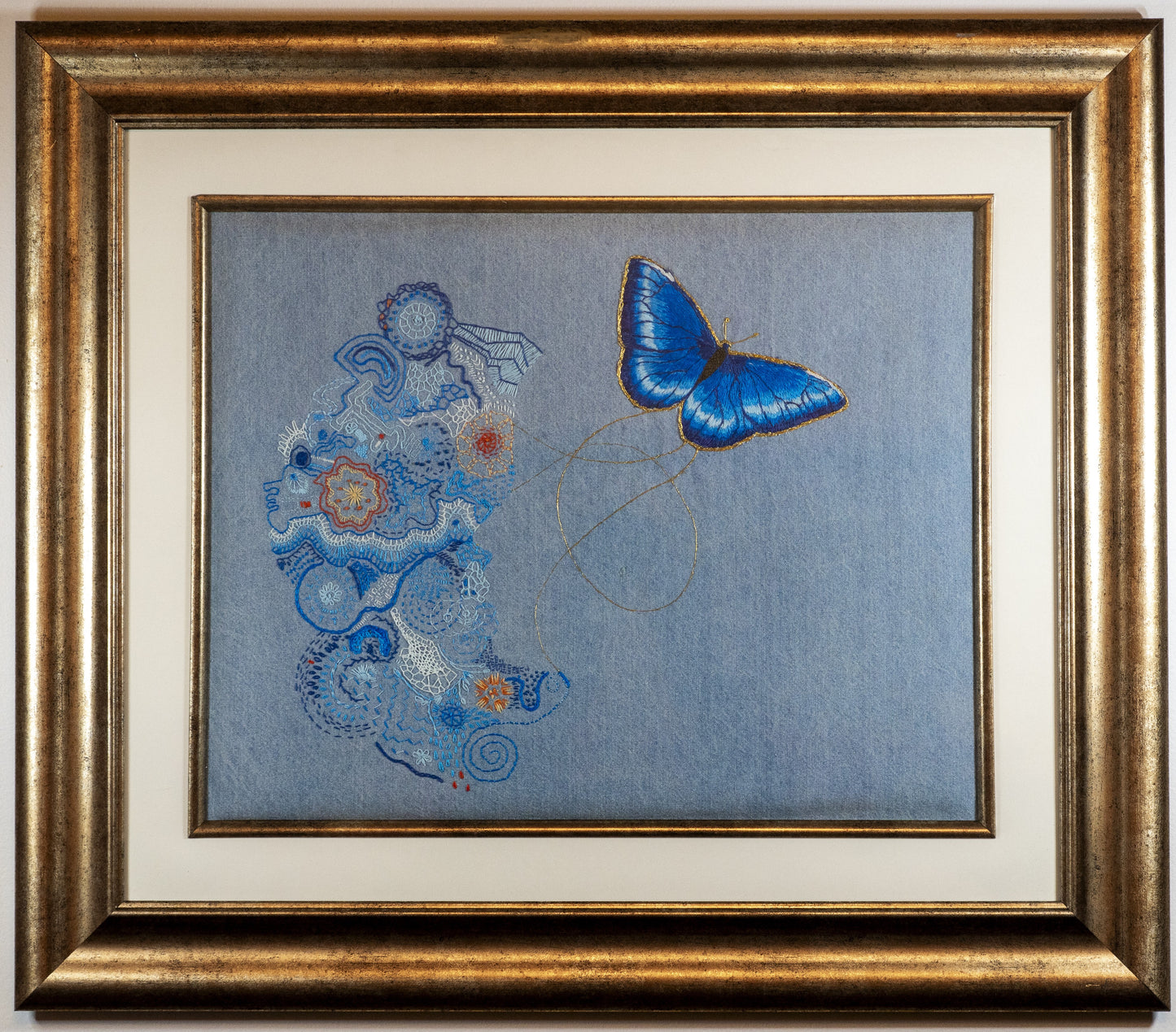 This is an embroidery work on denim in a gold frame. The image has a chaotic collection of organic looking embroidery stitches on the left. In the negative space where they stop is a profile. On the right is a detailed blue butterfly outlined n gold thread and joined to the left with golden threads. The overall feeling is of joy and light and colour.