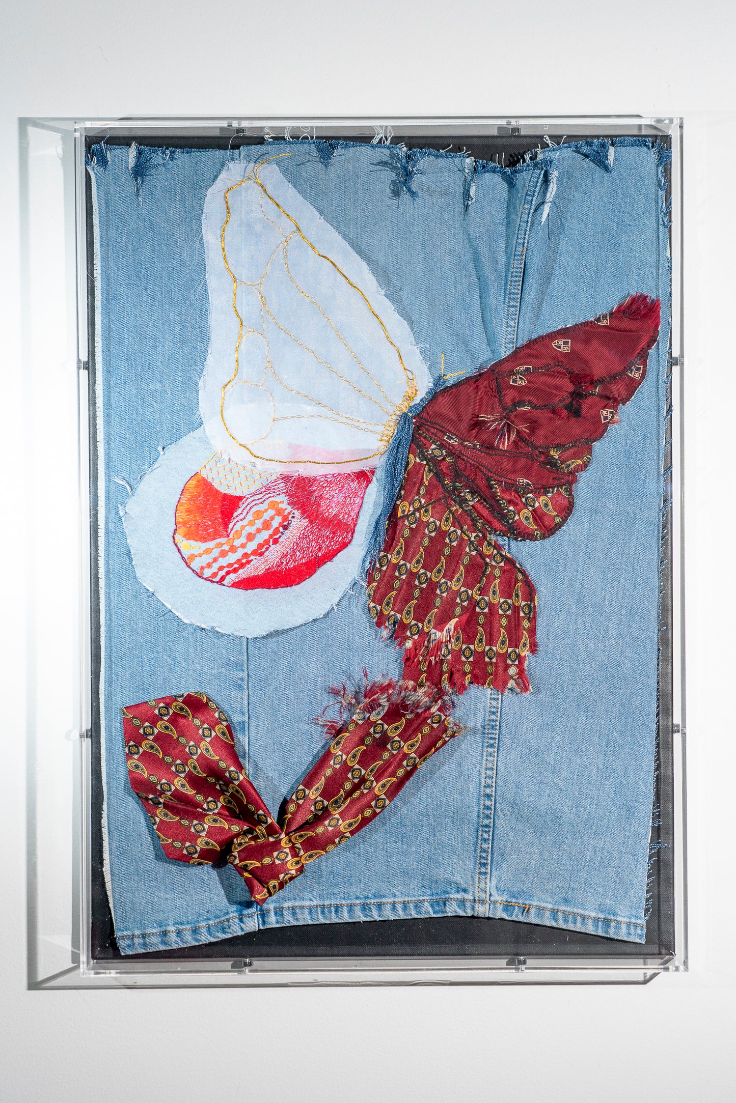 a large piece of reclaimed denim with the rolled seams visibly running thru it. at the bottom is a dark red business tie that has been shredded and reaches up to the butterfly emerging above it. The wing closest to the tie is still obviously sections of tie, the next up is more mosaic of tie pieces, the third is filled with colorful embroidery done on gauze appliqued to the denim and the top most wing is gauze the is outlined in gold thread that is fraying upwards into freedom