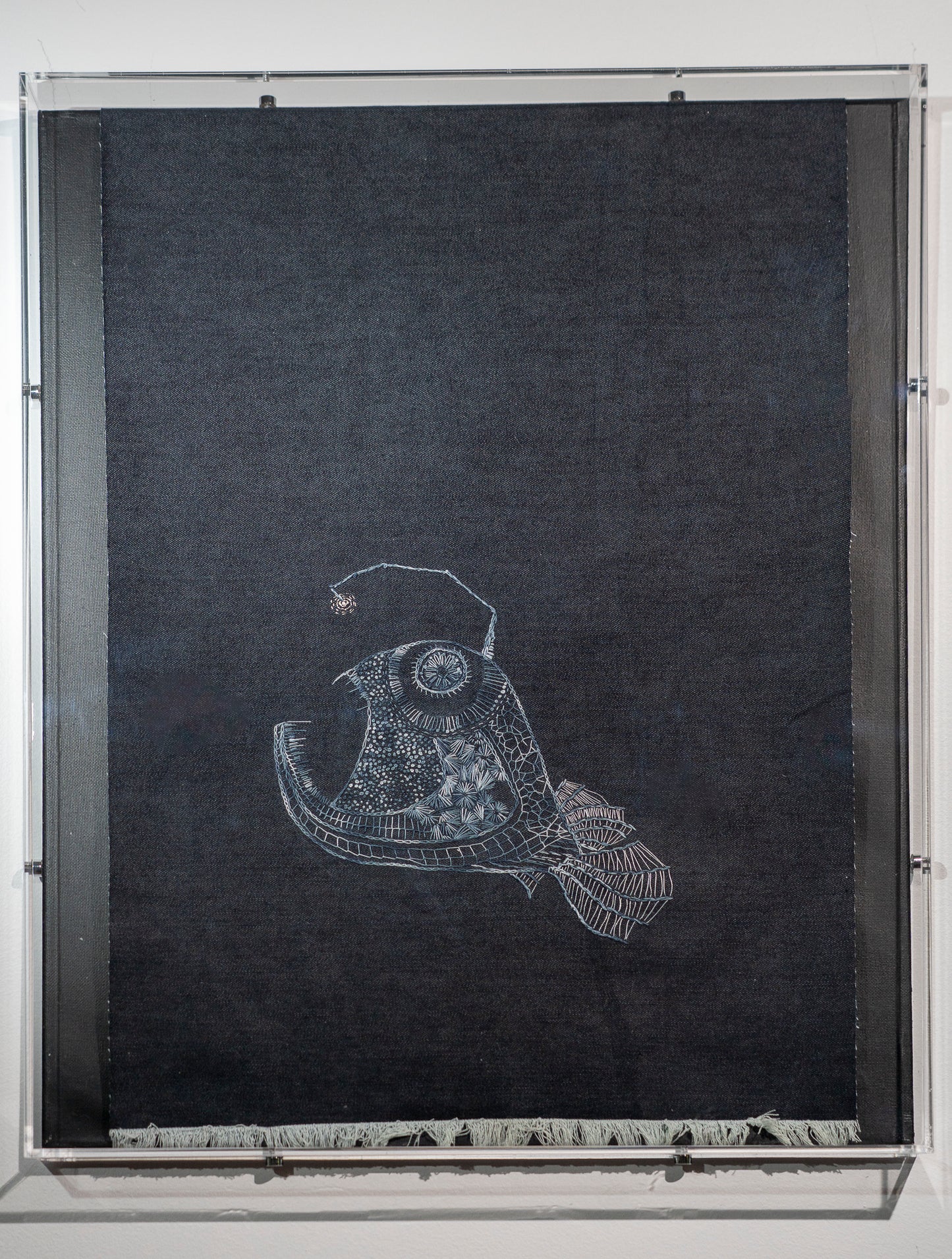 It is a large piece of dark denim with an angler fish embroidered in shades of grey in a contemporary embroidery style. She is inspired by the angler fish that was an internet sensation recently. Named it Hope. The top of the denim wraps into the frame so it is out of sight and the bottom is frayed.