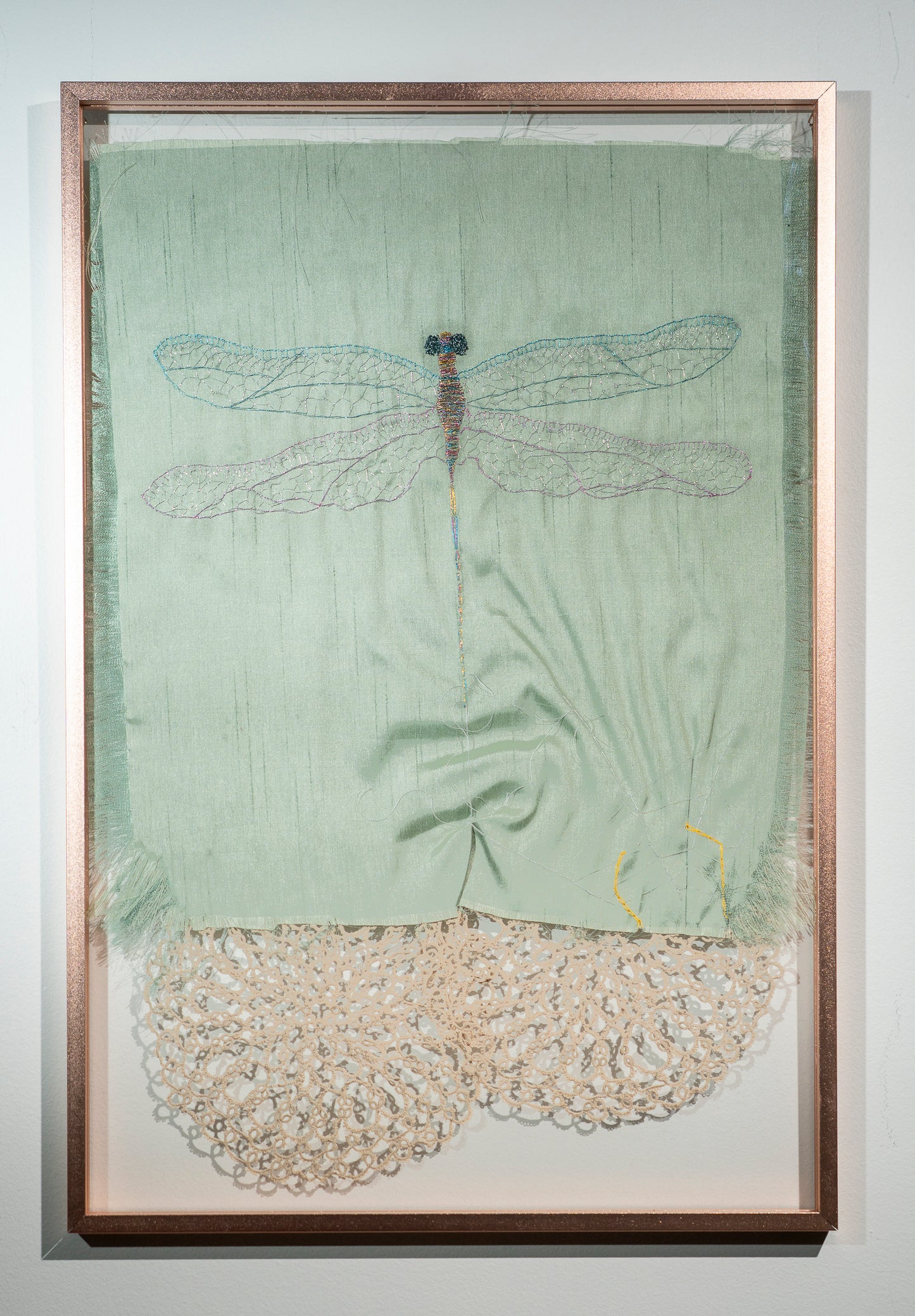 A dragonfly, done in iridescent thread, on raw green silk is escaping from the suggestion of a spider in a web. Vintage lace doilies peek out from below hinting at webs and insect eyes.