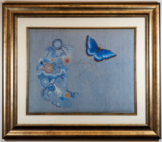 This is an embroidery work on denim in a gold frame. The image has a chaotic collection of organic looking embroidery stitches on the left. In the negative space where they stop is a profile. On the right is a detailed blue butterfly outlined n gold thread and joined to the left with golden threads. The overall feeling is of joy and light and colour.