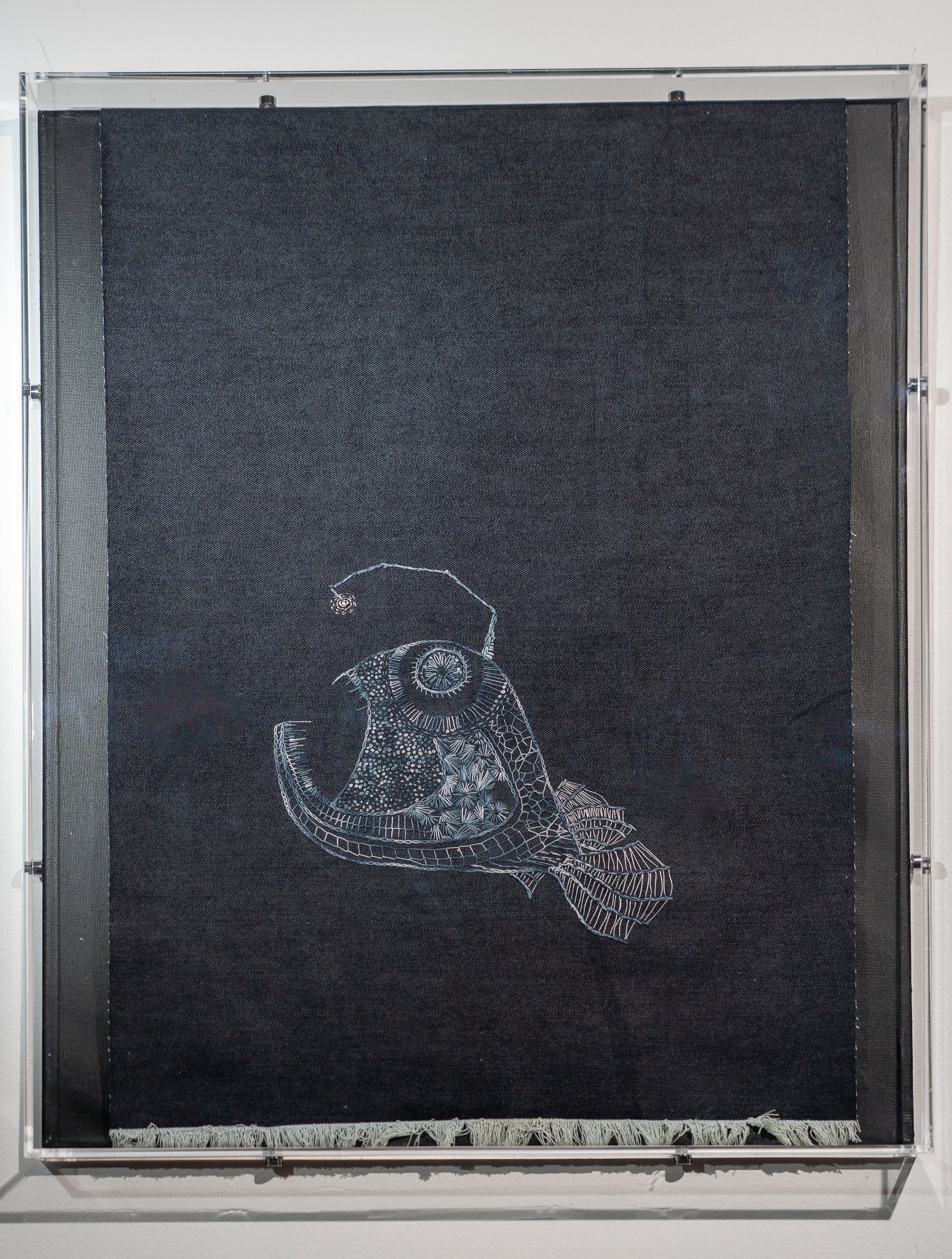 It is a large piece of dark denim with an angler fish embroidered in shades of grey in a contemporary embroidery style. She is inspired by the angler fish that was an internet sensation recently. Named it Hope. The top of the denim wraps into the frame so it is out of sight and the bottom is frayed.