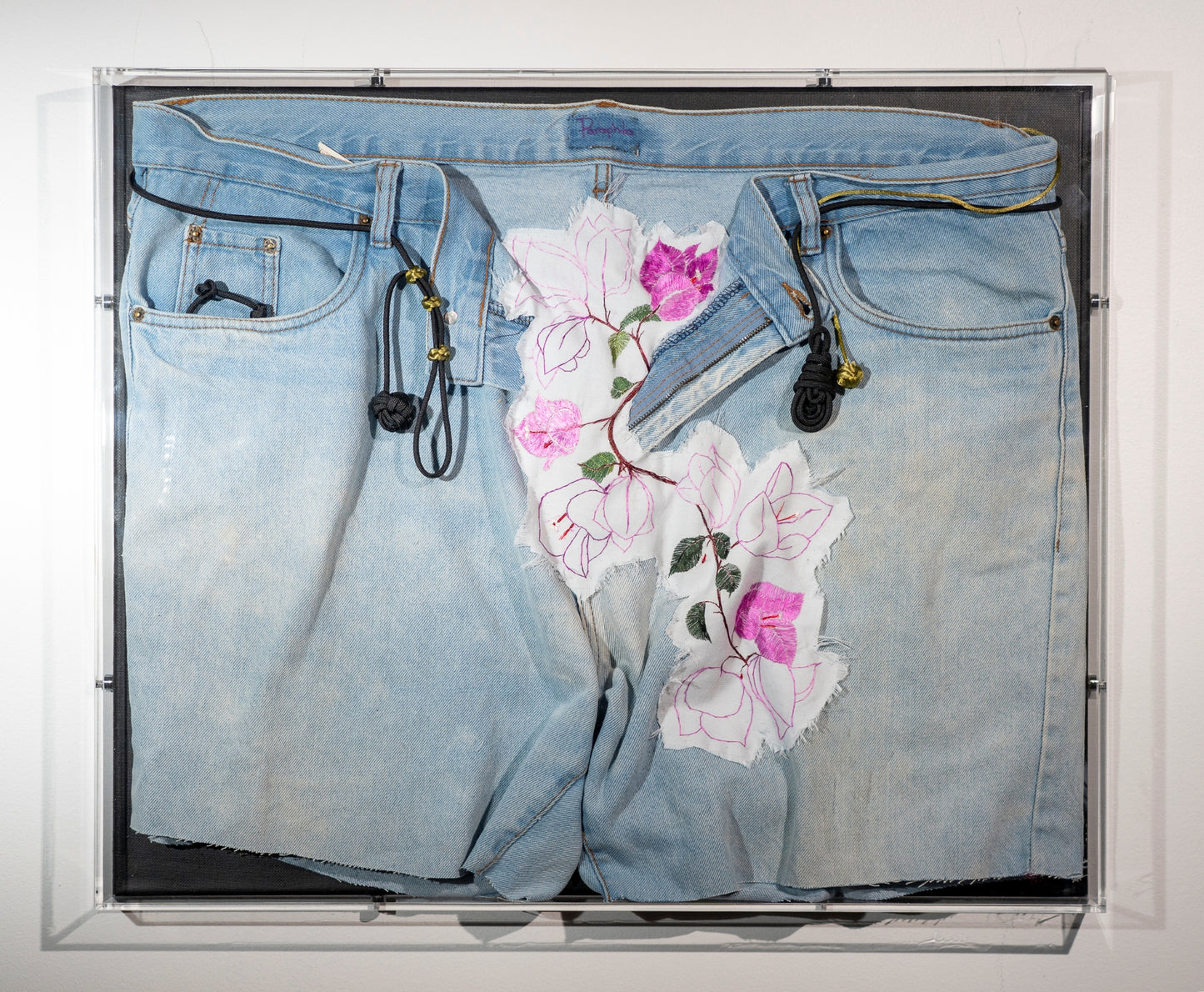 A pair of legless used jeans are displayed with bougainvillea vines erupting from the open zip. They are embroidered onto a frayed piece of cotton and worked into the fabric of the jeans. Through the belt loops are black and green cords that are tied in knots the include a monkeys fist, ranger beads and a pipa knot. The piece is hung in an acrylic display box.
