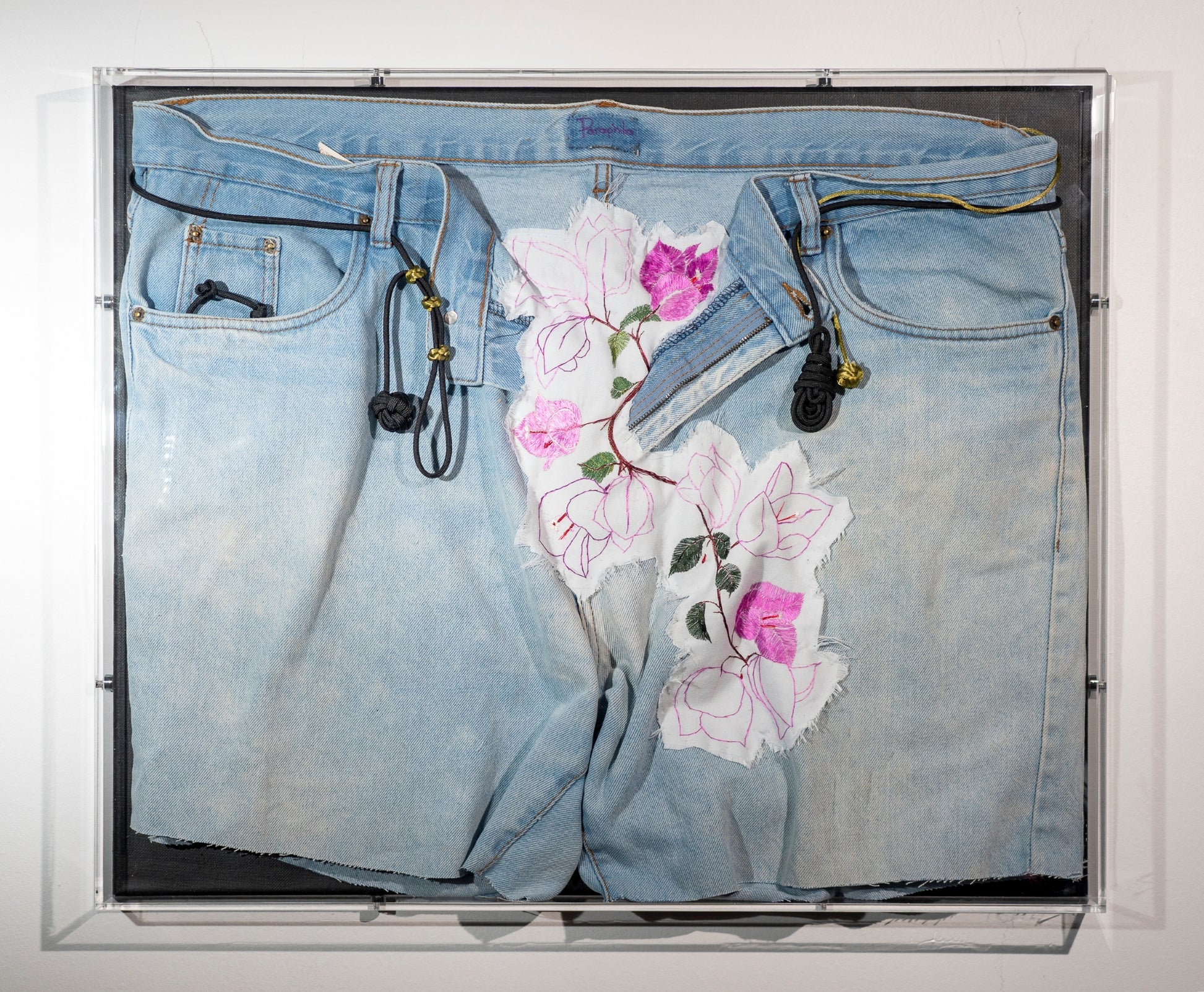 A pair of legless used jeans are displayed with bougainvillea vines erupting from the open zip. They are embroidered onto a frayed piece of cotton and worked into the fabric of the jeans. Through the belt loops are black and green cords that are tied in knots the include a monkeys fist, ranger beads and a pipa knot. The piece is hung in an acrylic display box.